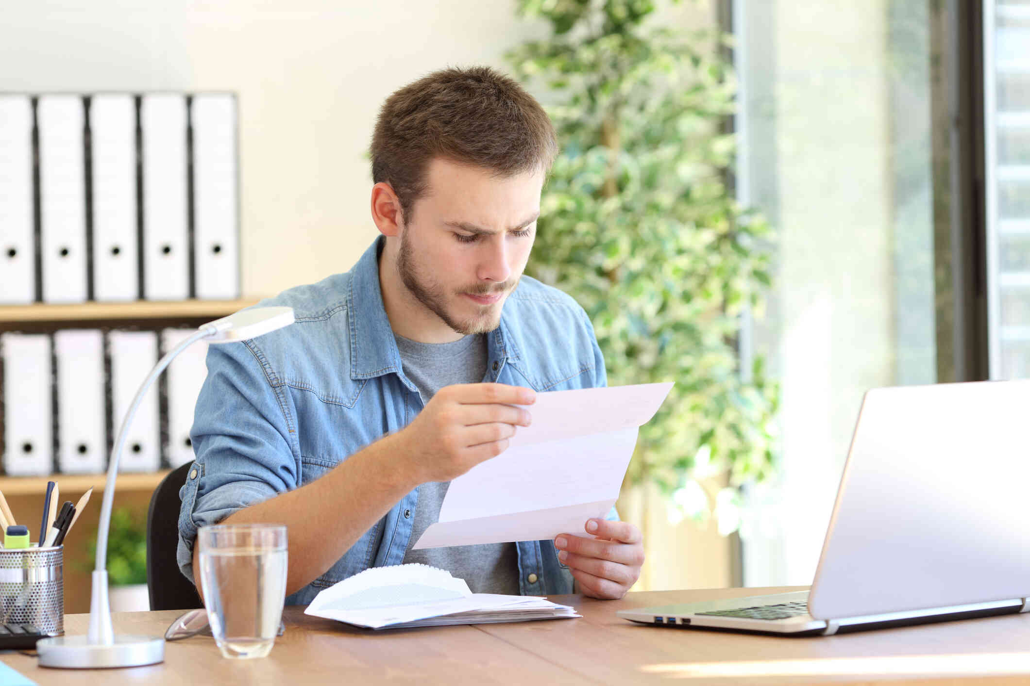 A man sits at a table in his home with his laptop open infront of hi mas he loks at a paper in his hand with a serious expression.